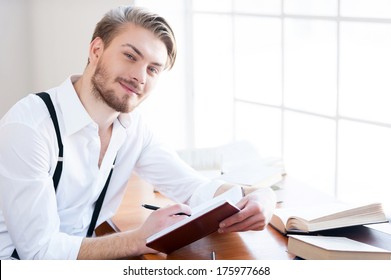 Inspired Author. Handsome Young Man In Shirt And Suspenders Writing Something In Note Pad And Smiling At Camera While Sitting At His Working Place