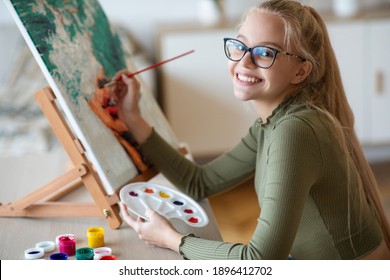 Inspired blonde teen girl with brush and palette painting, smiling at camera home interior. Creative schooler enjoying her new hobby, painting still life, side view. Teenagers lifestyle concept