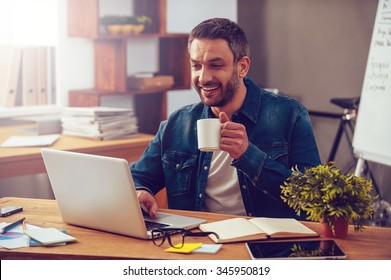 Inspired with cup of fresh coffee. Confident young man working on laptop and holding coffee cup while sitting at his working place in office - Powered by Shutterstock