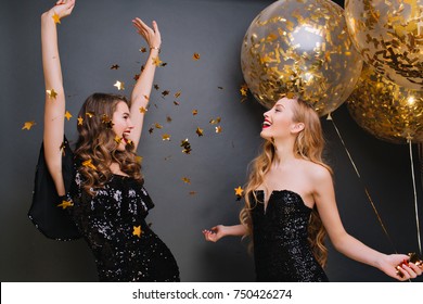 Inspired curly pale woman singing with hands up on dark background. Romantic blonde girl in black outfit holding party balloons and looking at friend which dancing under confetti. - Shutterstock ID 750426274