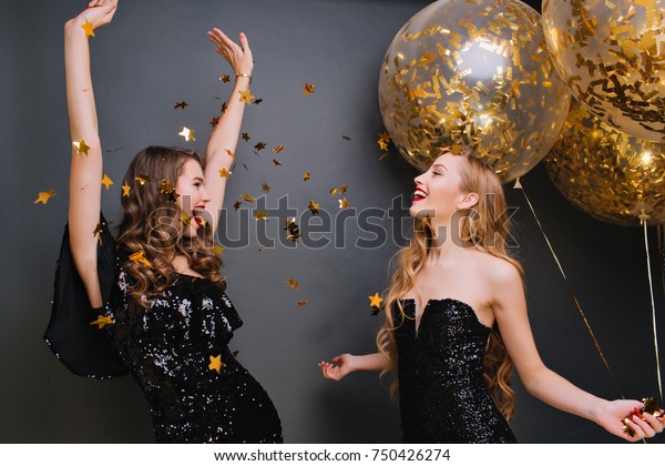 Inspired curly pale
woman singing with hands up on dark background. Romantic blonde
girl in black outfit holding party balloons and looking at friend
which dancing under
confetti.