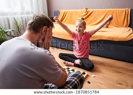 Inspired family. Happy girl-child holding her arm upwards while rejoicing her winning at the checkers game while playing with her father. Stock photo