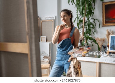 Inspired female artist reflects looking at painting. Smiling exited girl with palette put hand to chin evaluating work done thinking about next step. Dreamy contented woman painter at art studio. - Powered by Shutterstock