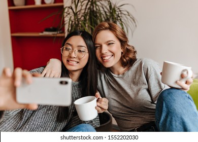 Inspired Girls Posing At Home With Coffee Cups. Cheerful Asian Woman Taking Selfie With Friends During Tea Time.