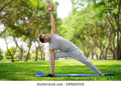 Inspired Indian Man Doing Yoga Asanas In City Park. Young Citizen Exercising Outside And Standing In Yoga Side Angle Pose. Fitness Outdoors And Life Balance Concept