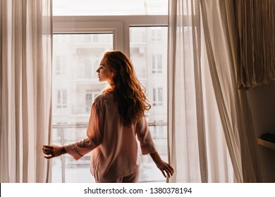 Inspired Long-haired Woman Posing In Pajama. Indoor Photo Of Serious Female Model Standing Near Window In Morning.