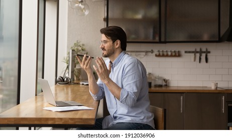 Inspired male remote worker sit at home office in kitchen before laptop pc dictate audio message via voice app on cell. Young man freelancer ask mobile phone ai assistant to search information online - Powered by Shutterstock