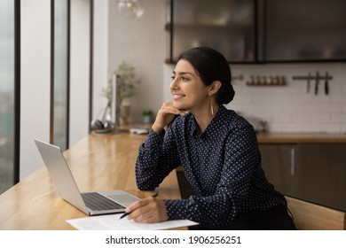 Inspired mixed race female sit by table with laptop at home kitchen look at window engaged by amazing idea. Dreamy young indian woman writer create new exciting story prepare to put it down on paper