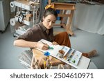 Inspired pensive woman artist sketching green plants with watercolors in notepad sitting on floor in creative workshop. Pleased girl freelance painter spends time with paintbrush, album in art studio.