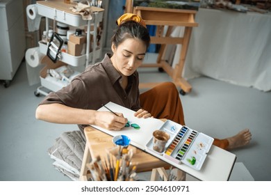 Inspired pensive woman artist sketching green plants with watercolors in notepad sitting on floor in creative workshop. Pleased girl freelance painter spends time with paintbrush, album in art studio. - Powered by Shutterstock