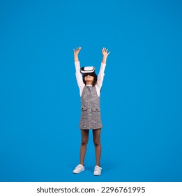 Inspired smiling black little kid in VR glasses plays online game, has fun, hands up isolated on blue background, studio, full length. Technology, childhood and emotions from virtual reality - Powered by Shutterstock