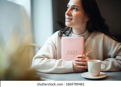 Inspired young woman with a smile looking through window and holding coral colored diary 2021. Hope and inspiration concept. Lady is smiling and dreaming about future new year. Happiness and success.