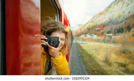 Inspiring female traveler and travel blogger look out of window of tourist train in cinematic beautiful mountains. Young beautiful woman hang out of window of red train. Excited world explorer