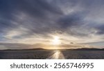 An inspiring sunset illuminates the West Coast of British Columbia with vibrant light, casting reflections over tranquil waters and depicting serene mountains in the distance against a cloudy sky.