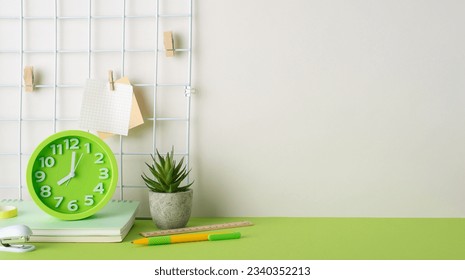 Inspiring workspace concept. Side view photo of grid notice board, clothespins, pen, ruler, tape, notepads, mini stapler, flowerpot, clock, arranged on green desk. Beige wall background for text or ad