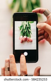 Instagram Photographer, Blogging Workshop Concept. Hand Holding Phone And Taking Photo Of Stylish Flower Flat Lay. Pink Tulips On White Wooden Rustic Background.space For Text