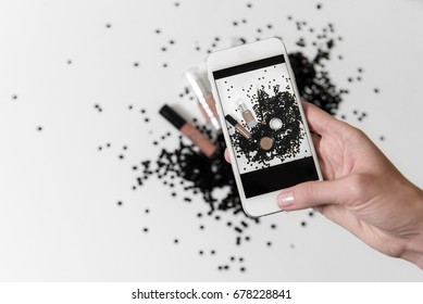 Instagram Photography Blogging Workshop Concept. A Girl Hanging A Phone Taking A Photo Of Cosmetics In A Black Paillette Mess On A White Background