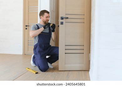 Installation of a lock on the front wooden entrance door. Portrait of young locksmith workman in blue uniform installing door knob. Professional repair service. Maintenance Concept. - Powered by Shutterstock