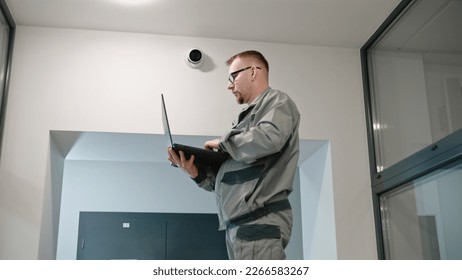 Installer in uniform sets up security camera in office room using laptop. Man in glasses checks CCTV cameras in computer program. Monitoring system. Concept of surveillance system and privacy.