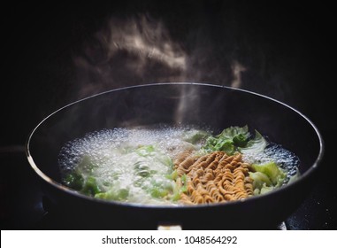 Instant noodles and green vegetables boiled in a pot. The boiling smoke on a black background.