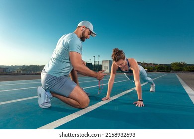 Instructor and athlete runner on the track. The athletics marks the time in the plank exercise on a stopwatch. Fitness trainer and mentee.