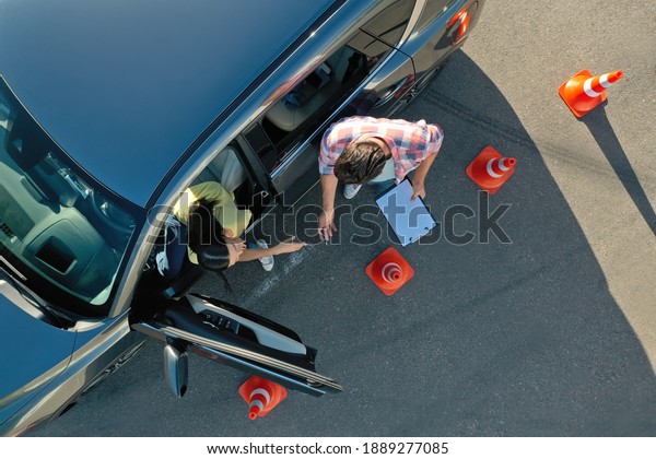 Instructor with clipboard near car\
and his student outdoors, above view. Driving school\
exam