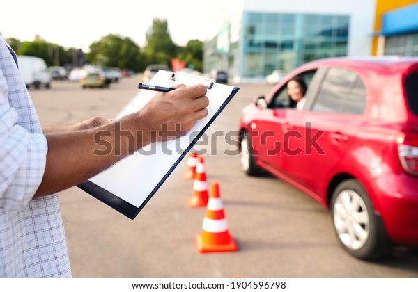 Instructor with clipboard near car outdoors,\
closeup. Driving school\
exam