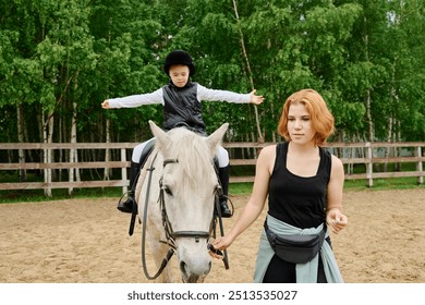 Instructor guiding young rider on white horse in outdoor equestrian arena surrounded by trees, with both focusing on their tasks, creating engaging equestrian experience for child - Powered by Shutterstock