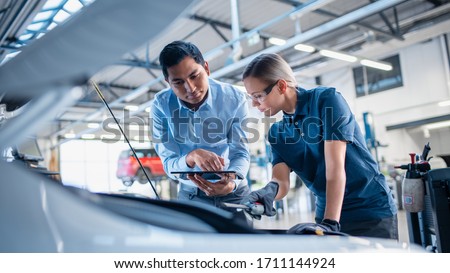 Instructor with a Tablet Computer is Giving a Task for a Future Mechanic. Female Student Inspects the Car Engine. Assistant is Checking the Cause of a Breakdown in the Vehicle in a Car Service.