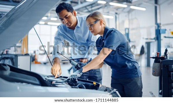 Instructor with a Tablet Computer is Giving a
Task for a Future Mechanic. Female Student Inspects the Car Engine.
Assistant is Checking the Cause of a Breakdown in the Vehicle in a
Car Service.