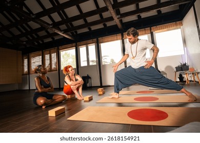 Instructor of yoga demonstrating a pose to two students in a serene studio with wooden beams and large windows.
