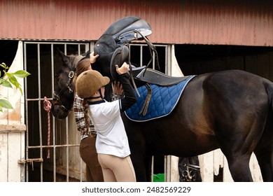 Instructor and young rider girl wearing safety helmet and polo shirt placing a saddle on a horse at a stable. - Powered by Shutterstock