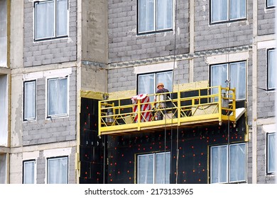 Insulation facade wall with mineral wool. Worker on suspended cradle, scaffolding platform for external works at height. Heat insulation of new building. Man on cradle facing wall with rock wool