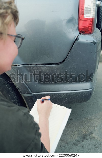 Insurance agent, middle-aged woman,\
conducts pre-insurance inspection of car. A woman makes notes in a\
notebook and fixes the damage on the car. Blurred\
foreground