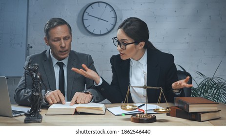 Insurance Agent Showing Shrug Gesture Near Colleague Reading Book, Laptop And Scales In Office 