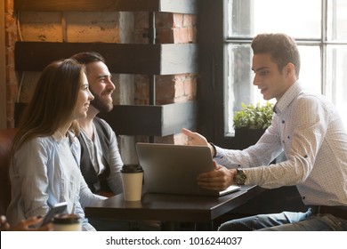 Insurance broker or salesman making offer to young millennial couple using laptop in cafe, realtor consulting customers about mortgage sitting at coffeehouse table pointing on computer screen