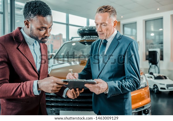 Insurance contract. Concentrated\
businessman signing a contract buying a car in a car\
showroom.