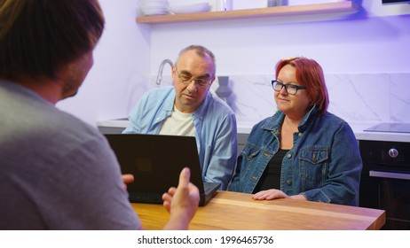 Insurer Man Presents Offer Medical Insurance Services To Aged Spouses Concept. Touristic Agency Worker Explain Showing On Laptop Travel Offers Tell About Tour Prices To Happy Older Couple In Office.