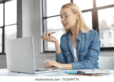 Intelligent blonde female uses voice command or recorder on smart phone in office works on laptop computer, surrounded with papers, sits at cabinet. Business, technology, communication concept - Powered by Shutterstock