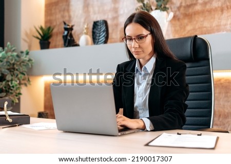 Intelligent caucasian beautiful brunette woman with glasses, in a formal suit, top manager, realtor, broker, sits at a desk in modern office, works in a laptop, chatting online with clients