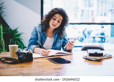Intelligent female student writing informative text to education textbook sitting at cafeteria table and using smartphone, millennial woman with mobile phone making notes preparing to exams - Powered by Shutterstock