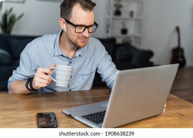 An Intelligent Guy Wearing Glasses And Smart Casual Shirt Sits At The Desk With A Cup Of Coffee And Using Laptop For Remote Work. A Young Man Develops Software, Works On Outsourcing