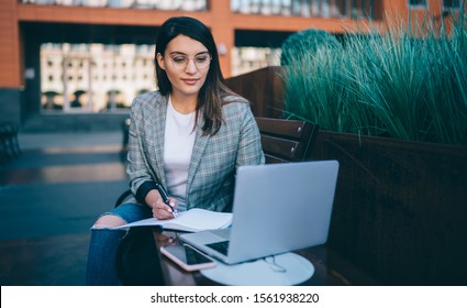 Intelligent lady in glasses making notes in planner and browsing laptop while sitting on bench in yard of modern building in city