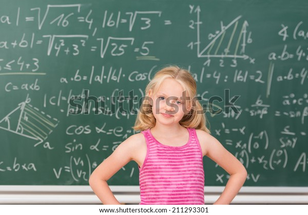 Intelligent little girl\
child prodigy in class standing confidently in front of a\
blackboard covered in mathematical equations with her hands on her\
hips smiling at the\
camera