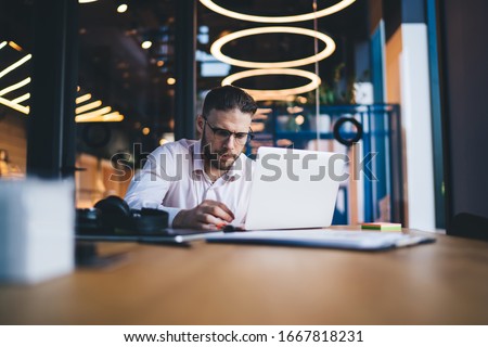 Intelligent male software developer in optical glasses typing program code during remote working on freelance, skilled businessman booking tutorial webinar on sale connecting to 4g wireless on laptop