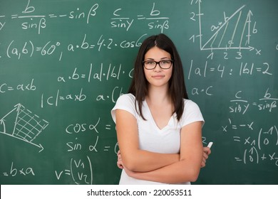 Intelligent young female maths student or teacher wearing glasses standing in front of a chalkboard with mathematical equations with folded arms and a piece of chalk in her hand - Powered by Shutterstock