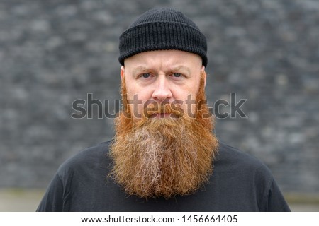 Intense man with bushy red beard wearing a beanie hat scowling at the camera with a penetrating stare over grey