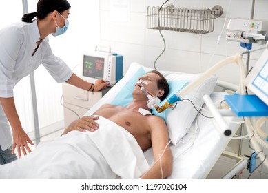Intensive care unit. Young lady in white lab coat checking digital device for measuring blood pressure. Middle aged man with endotracheal tube in mouth sleeping after surgery