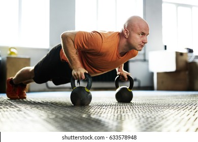 Intensive training in modern gym illuminated with bright sunlight: bald middle-aged sportsman standing in plank position with help of kettlebells, full-length portrait - Powered by Shutterstock