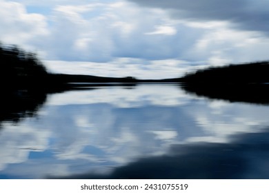 Intentional camera movement (ICM) image of a dream like lake scene with dark forest and cloudy spring sky reflectiong on water surface created by motion blur.